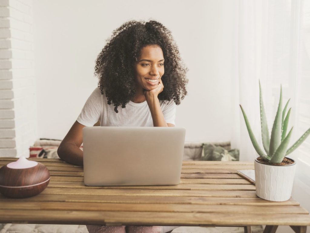 Woman engaging in an online somatic therapy session from her Los Angeles home. Woman is smiling with a laptop on a wooden desk.