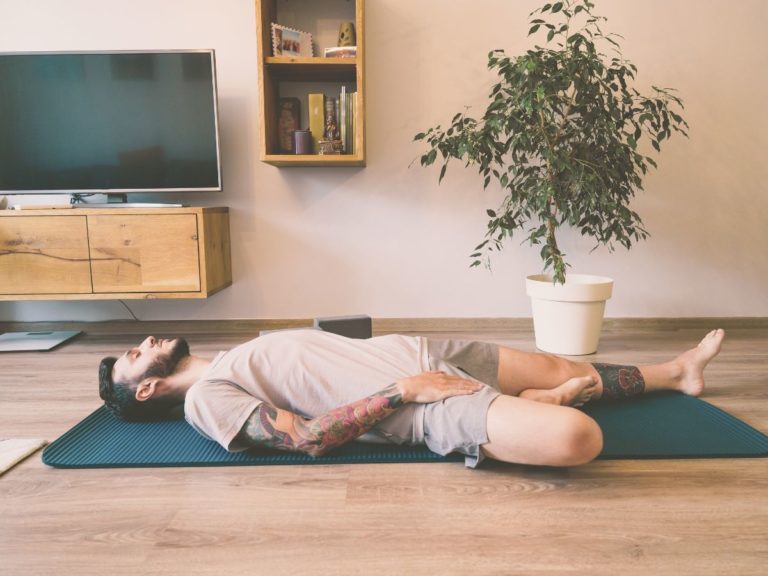 Man laying on a mat in a somatic therapy office in Los Angeles. Man has tattoos and is wearing a tan shirt and tan shorts. Office is clean and nice. Hardwood floors.