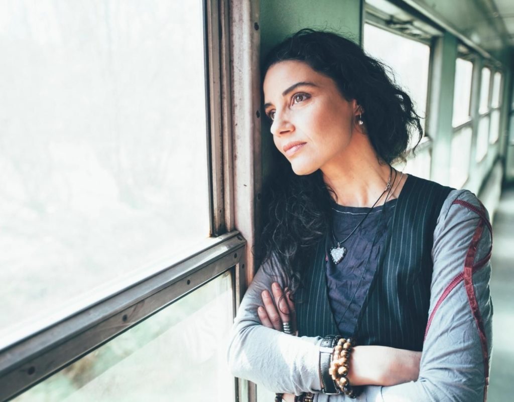 Woman with black hair looking out window, wondering what to expect in therapy. Woman wears black shirt and bracelet.