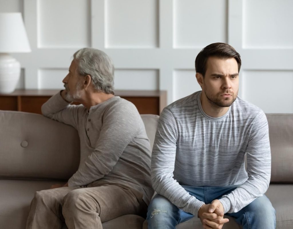 Adult Child of Narcissistic parent: narcissistic father sitting with son, both are cold and distant, not looking at each other on brown couch. both wearing gray long sleeves, white background.