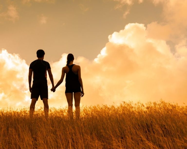 A couple in Los Angeles walking to trauma therapy. Couple is holding hands and walking on brown grass. Clouds and sunset in background.