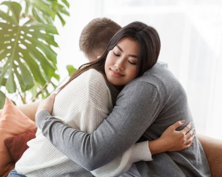 A couple embracing a hug in attachment therapy session in LA, showing that communication in therapy can help couples and relationships. Couch is gray, both people wearing long sleeve shirts, and look content with each other.