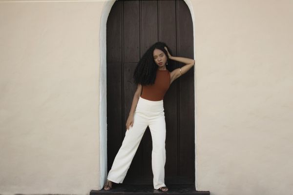 Woman standing in the doorway of her Los Angeles apartment, reflecting personal reflection and growth through attachment therapy. Woman is wearing white pants, brownshirt, and leaning on a brown door.