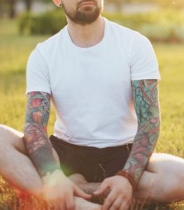 Man with tattoos doing somatic therapy work in Los Angeles park. He is sitting criss cross style on green grass.